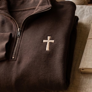 May include: A dark brown quarter-zip sweatshirt featuring a small, embroidered cream-colored cross on the left chest. The garment has a high collar with a metal zipper closure and ribbed cuffs, showcasing a simple and clean design aesthetic.