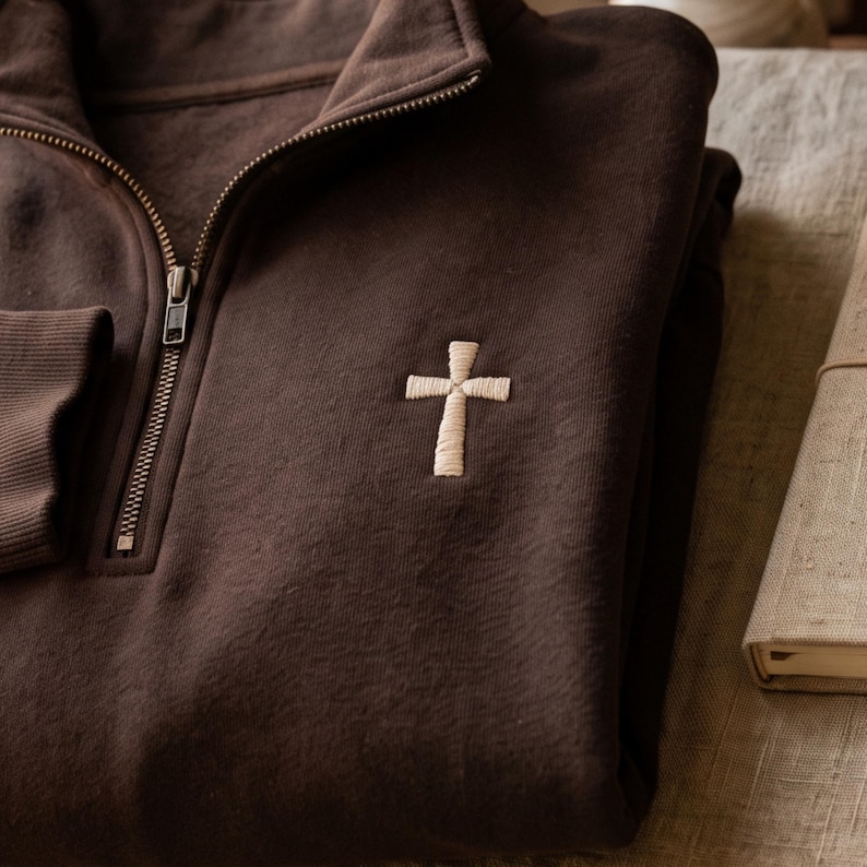 May include: A dark brown quarter-zip sweatshirt featuring a small, embroidered cream-colored cross on the left chest. The garment has a high collar with a metal zipper closure and ribbed cuffs, showcasing a simple and clean design aesthetic.