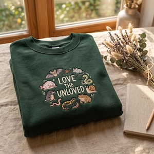 Peut inclure: Un sweat-shirt à col rond vert foncé avec un motif circulaire brodé représentant divers animaux dont une chauve-souris, une araignée, une souris, un serpent, un escargot, un crapaud, une salamandre, un ver, une taupe et un blobfish.