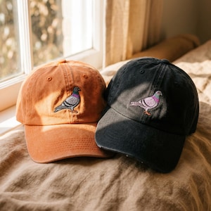 Puede incluir: Dos gorras de béisbol, una en color naranja apagado y otra en negro desteñido, cada una con un diseño bordado de una paloma en el panel frontal. Las gorras tienen una corona estructurada, una visera curva y una construcción clásica de seis paneles.
