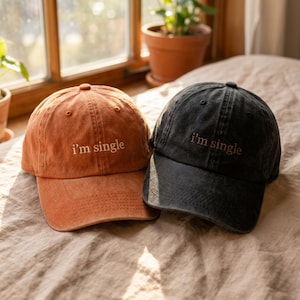 Könnte beinhalten: Zwei Baseballkappen, eine in verwaschenem Orange und eine in verwaschenem Schwarz, beide mit dem gestickten Text 'i'm single' in einer einfachen Schriftart auf der Vorderseite. Jede Kappe hat einen gebogenen Schirm und ein sechsteiliges Design.
