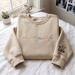 Puede incluir: Una sudadera beige de cuello redondo con el texto bordado 'it's a good day to X-ray' en colores pastel con pequeñas estrellas doradas. La manga muestra un gráfico negro de un esqueleto saludando sobre el nombre 'Ashley' en letras negras.