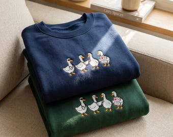 May include: A navy blue and a dark green crewneck sweatshirt, each featuring an embroidered graphic of four white ducks dressed as medical professionals, including one with a stethoscope and one holding a clipboard with a red heart icon.
