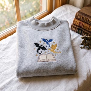 May include: A light gray crewneck sweatshirt featuring an embroidered design of an open book with three small dragons in black, blue, and gold flying above it, surrounded by small embroidered stars.