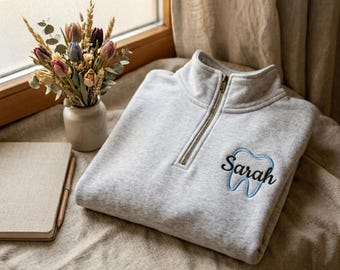 May include: A light gray quarter-zip sweatshirt featuring an embroidered design on the left chest. The embroidery consists of a light blue tooth outline with the name Sarah stitched in black cursive script across the center of the tooth.