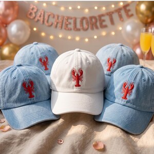 Può includere: Una collezione di cappellini da baseball con un'aragosta rossa ricamata sul davanti. Un cappellino è bianco tinta unita, mentre gli altri sono in denim azzurro. Ogni cappellino ha una visiera curva, un bottone sulla parte superiore e occhielli.