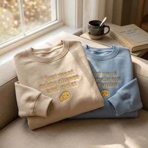 May include: Two crewneck sweatshirts, one in beige and one in light blue, each featuring the embroidered text "I just want some cheese and quiet" in gold thread, accompanied by a small embroidered yellow Swiss cheese wedge graphic on the front.