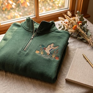 May include: A forest green quarter-zip sweatshirt featuring an embroidered mallard duck in flight over water with reeds and autumn leaves. The garment has a metal zipper closure at the collar and is crafted from a soft, solid-colored fabric.
