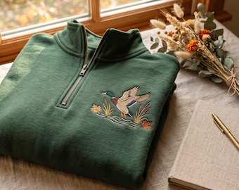 May include: A forest green quarter-zip sweatshirt featuring an embroidered mallard duck in flight over water with reeds and autumn leaves. The garment has a metal zipper closure at the collar and is crafted from a soft, solid-colored fabric.