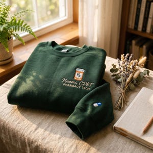 May include: A dark green crewneck sweatshirt featuring an embroidered orange prescription pill bottle icon above the text "Naomi, CPhT PHARMACY TECH" in white. The sleeve cuff displays a small embroidered blue and white capsule pill design.
