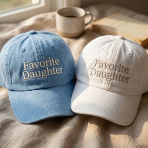 Może przedstawiać: Dwie czapki z daszkiem, jedna jasnoniebieska i jedna biała, każda z wyhaftowanym napisem "Favorite Daughter" w neutralnym odcieniu. Obie czapki mają usztywnianą koronę, zakrzywiony daszek i guzik na g&oacute;rze, ustawione obok siebie.