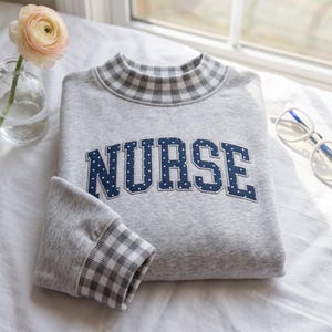 Może przedstawiać: Jasnoszara bluza z golfem oraz mankietami w szaro-białą kratkę typu gingham. Słowo NURSE zostało naszyte na piersi z ciemnoniebieskiego materiału w białe kropki, z wyraźnym białym obramowaniem wokół każdej z liter.