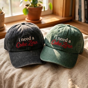 Op de afbeelding: Twee baseballpetten, één in antracietgrijs en één in bosgroen, beide voorzien van de geborduurde witte tekst i need a en de rode cursieve tekst Coke Zero. Elke pet heeft een gestructureerde kroon, een gebogen klep en stikseldetails.