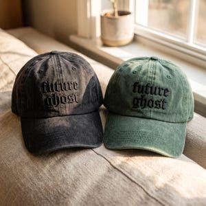 May include: Two baseball caps with a distressed finish, one in charcoal gray and one in sage green. Both caps feature the text "future ghost" embroidered in black gothic-style lettering on the front panel, with a curved brim and eyelets.