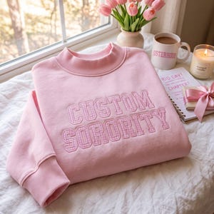 Peut inclure: Un sweat-shirt à col rond rose clair avec le texte CUSTOM SORORITY brodé dans un motif vichy rose et blanc. Le sweat-shirt présente un col montant côtelé et des poignets côtelés, offrant un design épuré et structuré pour une tenue décontractée.