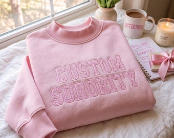 Puede incluir: Una sudadera de manga larga en color rosa claro con cuello alto y puños acanalados. El frente muestra el texto CUSTOM SORORITY en letras de bloque con un relleno de patrón de cuadros vichy rosa y blanco, bordado en la tela.