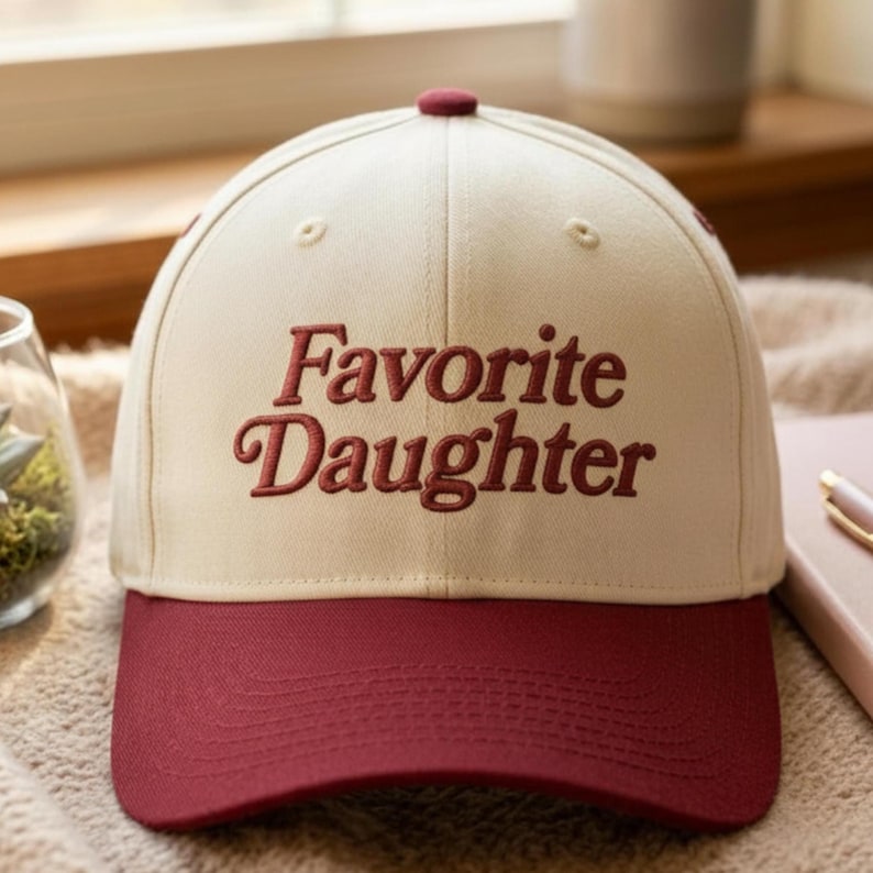 Puede incluir: Una gorra de b&eacute;isbol con corona de color crema y visera de color rojo oscuro. La parte delantera de la gorra presenta el texto bordado "Favorite Daughter" en una fuente serif de color rojo oscuro. La gorra tiene un bot&oacute;n a juego.