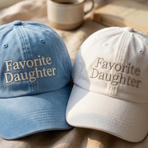 Może przedstawiać: Dwie czapki z daszkiem, jedna jasnoniebieska i jedna biała, każda z wyhaftowanym napisem "Favorite Daughter" czcionką szeryfową na przednim panelu. Obie czapki mają usztywnianą koronę, zakrzywiony daszek i guzik na środku u g&oacute;ry.
