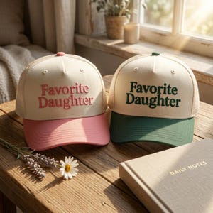 Puede incluir: Dos gorras tipo camionero con paneles frontales color crema y parte trasera de malla. La gorra izquierda tiene bordado rosa con el texto Favorite Daughter y visera rosa. La derecha tiene bordado verde oscuro Favorite Daughter y visera verde.