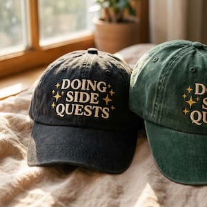 May include: A black baseball cap and a dark green baseball cap, both featuring embroidered text that reads DOING SIDE QUESTS in a light cream colour, accented by small four-pointed star designs in a golden yellow thread on the front panels.