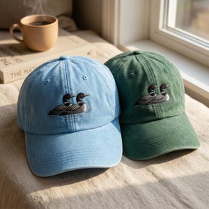 Könnte beinhalten: Zwei Baseballkappen, eine hellblau und eine dunkelgrün, jeweils mit einer Stickerei von zwei auf dem Wasser schwimmenden Eistauchern. Die Kappen haben eine strukturierte Krone, einen gebogenen Schirm und genähte Ösen zur Belüftung.