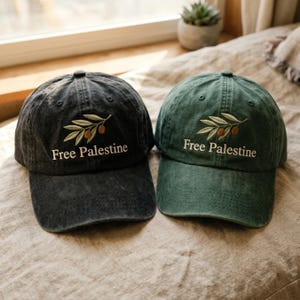 以下が含まれることがあります： チャコールグレーとフォレストグリーンの野球帽が2つ。どちらも前面中央にオリーブの枝の刺繍と白い文字で「Free Palestine」と記されています。各キャップはカーブしたつばと6枚パネル構造が特徴です。