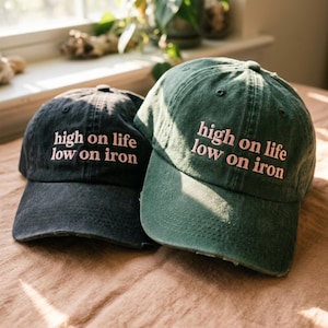 May include: Two baseball caps, one in charcoal black and one in forest green, both featuring the embroidered text 'high on life low on iron' in light pink thread. Each cap has a six-panel construction with a curved brim and a distressed finish.