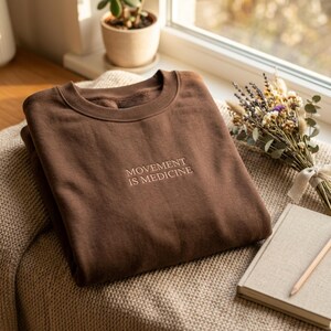 May include: A dark brown crewneck sweatshirt featuring the embroidered text "MOVEMENT IS MEDICINE" in a light beige thread centered on the chest. The garment has a ribbed collar and long sleeves, showcasing a simple and clean design.