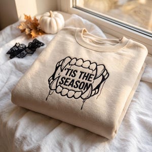 Pode incluir: Uma camisola bege de gola redonda com bordado preto de presas de vampiro rodeando o texto 'TIS THE SEASON. O design inclui dentes afiados e detalhes de gotejamento, centrados no peito da peça, feita de um tecido macio.