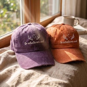 May include: Two baseball caps, one purple and one orange, featuring a white graphic of deer antlers and stars above the text "to whatever end" embroidered on the front. Both caps have a distressed, washed-out fabric texture and a curved brim.