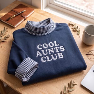 May include: A navy blue long-sleeve sweatshirt featuring a mock neck collar and cuffs with a blue and white gingham pattern. The front of the sweatshirt displays the white embroidered text COOL AUNTS CLUB in a bold, sans-serif font.