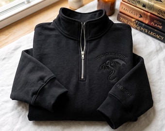 May include: A black quarter-zip sweatshirt featuring a silver metal zipper and a mock neck collar. The chest displays an embroidered dragon graphic with the text WINGLEADER above it, while the sleeve cuff features the embroidered text RIORSON.