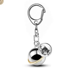 Puede incluir: Un llavero plateado con un dije redondo y pulido y un dije circular más pequeño. El dije más pequeño presenta una ilustración de gato y el nombre "Bob". El dije más grande tiene una banda dorada alrededor de su centro.