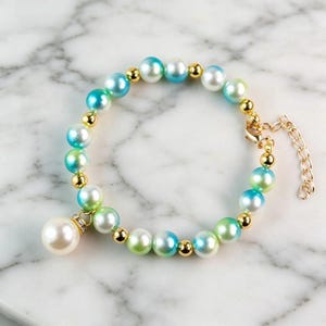 May include: A bracelet featuring alternating round beads in shades of blue, green, and white, interspersed with small gold-toned metallic spheres. A larger white pearl-like bead hangs as a charm from the bracelet, which includes a gold-toned chain.
