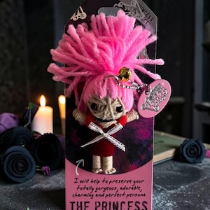 Op de afbeelding: Een kleine touwpop met felroze garen haar, zwarte kraaloogjes en een rode jurk met een wit lintkruis. De pop is bevestigd aan een roze kaart met de tekst "I will help to preserve your totally gorgeous, adorable, charming and perfect persona" en "THE PRINCESS".