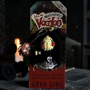 May include: A Watchover Voodoo Goth Girl doll with black yarn hair, a cream-colored string-wrapped face, red bead eyes, and a black outfit featuring a skull graphic and a plaid skirt. The doll is attached to a cardboard backing with the text Being Different Is Good and I will help you to not just be yourself, but your best self.