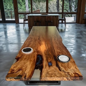 May include: A large, custom-made wooden dining table with a live edge design. The table features a rich, natural wood grain with a glossy finish. It has two built-in recessed areas and a metal base. A plate sits on the table.