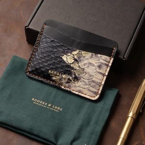 May include: A leather card holder featuring a black and gold snakeskin-textured panel, a dark green fabric pouch with the text RHODES & LORE, and a metallic gold pen. The card holder is embossed with the brand name RHODES & LORE in gold lettering.