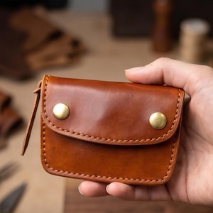 May include: A small, rectangular brown leather wallet featuring a flap closure secured by two round brass-colored metal snaps. The wallet is constructed with visible stitching along the edges and includes a leather zipper pull on the side.
