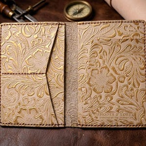May include: A beige leather wallet featuring an intricate embossed floral pattern with gold-coloured highlights. The interior includes multiple card slots and a central pocket. The text RHODES & LORE ENRICHED BY THE JOURNEY is stamped in the corner.