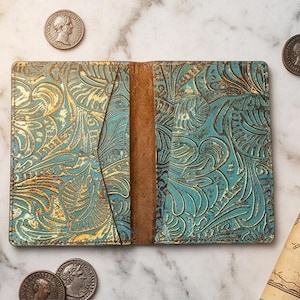 May include: A rectangular leather passport holder featuring a teal and gold embossed floral pattern with a central brown spine. The item is displayed alongside a brass compass, several antique-style metal coins, and a rolled parchment map.