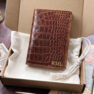 May include: A brown leather wallet with a crocodile skin texture, personalized with the initials "RML" in gold. The wallet is presented in a cardboard box with a white fabric lining and a white rope drawstring.