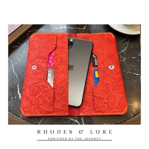 Op de afbeelding: Een felrode lederen portemonnee-hoes met een reliëf bloemenpatroon, gele stiksels en meerdere kaartsleuven. Een donkergrijze smartphone is in het centrale vak geplaatst. De tekst RHODES & LORE ENRICHED BY THE JOURNEY is zichtbaar.