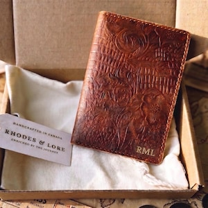 May include: A brown leather passport holder with an embossed floral and grid pattern, featuring the initials RML in the bottom right corner. A white tag attached to the item displays the text Handcrafted in Canada, Rhodes & Lore, Enriched by the Journey.