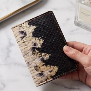 May include: A rectangular passport holder featuring a textured, scale-like pattern. The design transitions from a natural, light beige and cream color on the left side to a solid, deep black on the right side, with visible stitching along the edges.