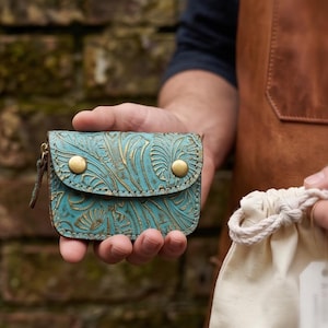 Könnte beinhalten: Eine kleine, türkisfarbene Lederbrieftasche mit floralem Prägemuster und goldfarbenen Knopfverschlüssen. Die Brieftasche hat einen Reißverschluss und wird in der Hand einer Person gehalten. Ein Kordelzugbeutel ist im Hintergrund.