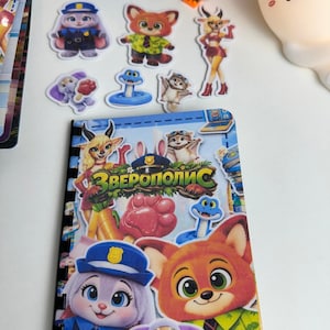 Op de afbeelding: Een Zootopia-thema notitieboek met een spiraalbinding, omringd door karakterstickers en blokfiguren. De kaft van het notitieboek bevat stripfiguren en de Cyrillische tekst "ЗВЕРОПОЛИС". Stickers bevatten een politiekonijn, een vos en andere dieren.