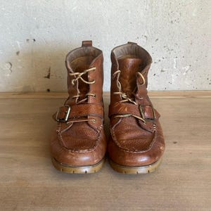 May include: A pair of brown leather ankle boots featuring a moccasin-style toe, lace-up front closure, and a single adjustable strap with a metal buckle across the vamp. The boots have a textured leather finish and a low, flat rubber sole.