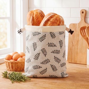 Può includere: Un sacco per il pane beige con un motivo a foglie nere, contenente due pagnotte. Un piccolo cestino di uova e un mazzetto di erbe sono nelle vicinanze. La borsa ha una chiusura a fibbia e si trova su una superficie di legno.