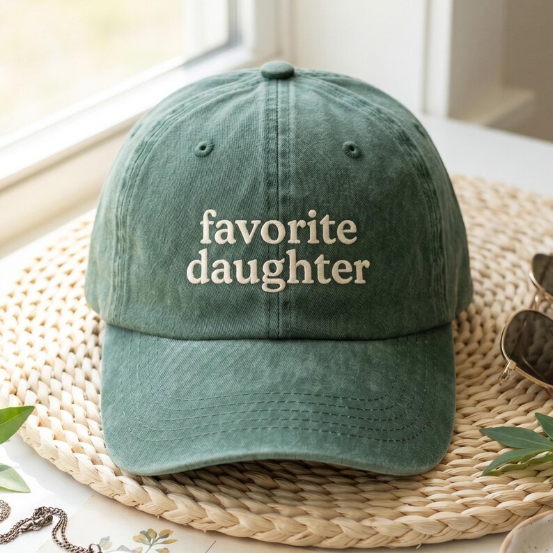 Pode incluir: Bon&eacute; de beisebol verde com o texto bordado "favorite daughter" em branco. O bon&eacute; &eacute; feito de um tecido macio e texturizado e tem uma aba curva. O chap&eacute;u est&aacute; sobre uma esteira tecida.